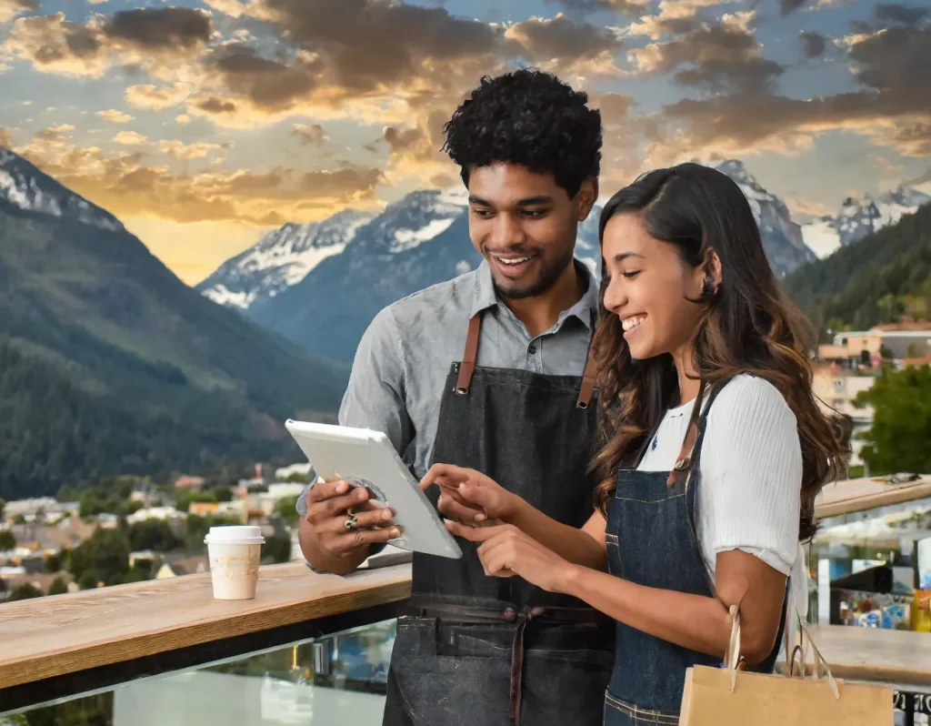 Two people in aprons using a tablet point of sale system outside, with mountains in the background and a coffee cup nearby.
